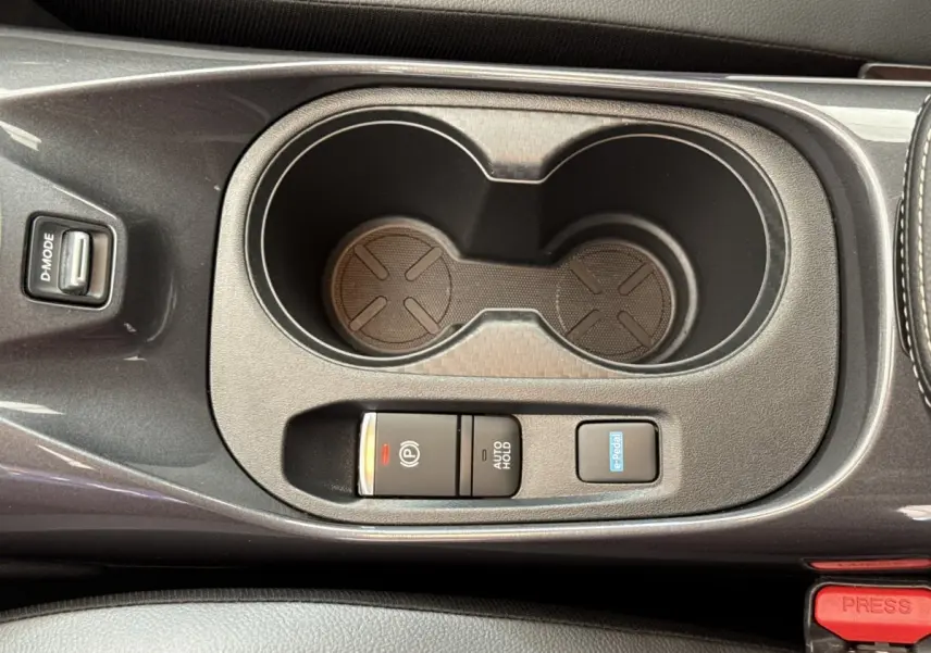 Vue rapprochée de la console centrale grise foncée du Nissan Juke 2023, avec porte-gobelets et boutons de frein électrique et e-Pedal.