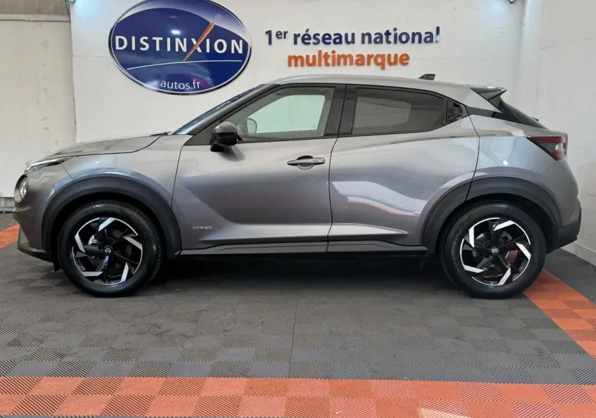 Vue de profil côté gauche d'un Nissan Juke gris foncé hybride 2023 avec jantes noires et toit noir contrasté.
