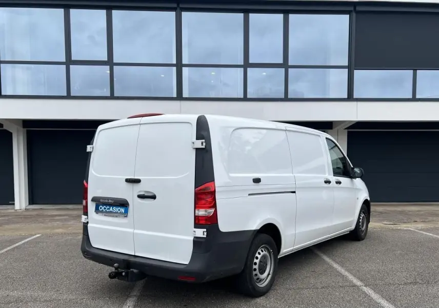 Vue 3/4 arrière droite d’un Mercedes Vito Fourgon blanc avec attelage et porte latérale, stationné devant un bâtiment moderne.