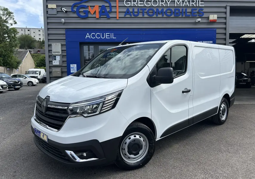 Renault Trafic Fourgon blanc en 3/4 avant droit, avec calandre noire et projecteurs modernes, stationné devant un garage.