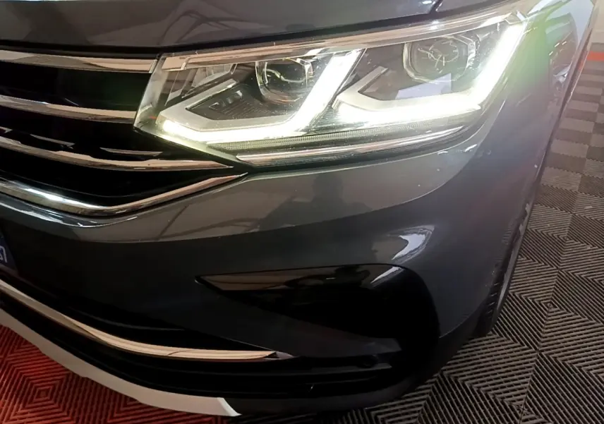 Gros plan sur le phare avant droit allumé d'un Volkswagen Tiguan gris clair avec calandre chromée visible.