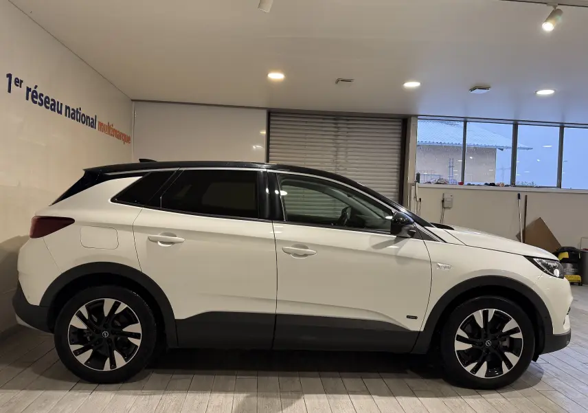 Vue latérale droite d’un Opel Grandland X blanc hybride rechargeable avec toit noir et jantes bicolores en intérieur showroom.