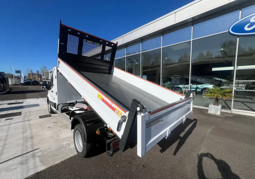 Vue 3/4 arrière droite d’un Ford Transit Chassis Cabine blanc avec benne basculante relevée devant un bâtiment Ford.