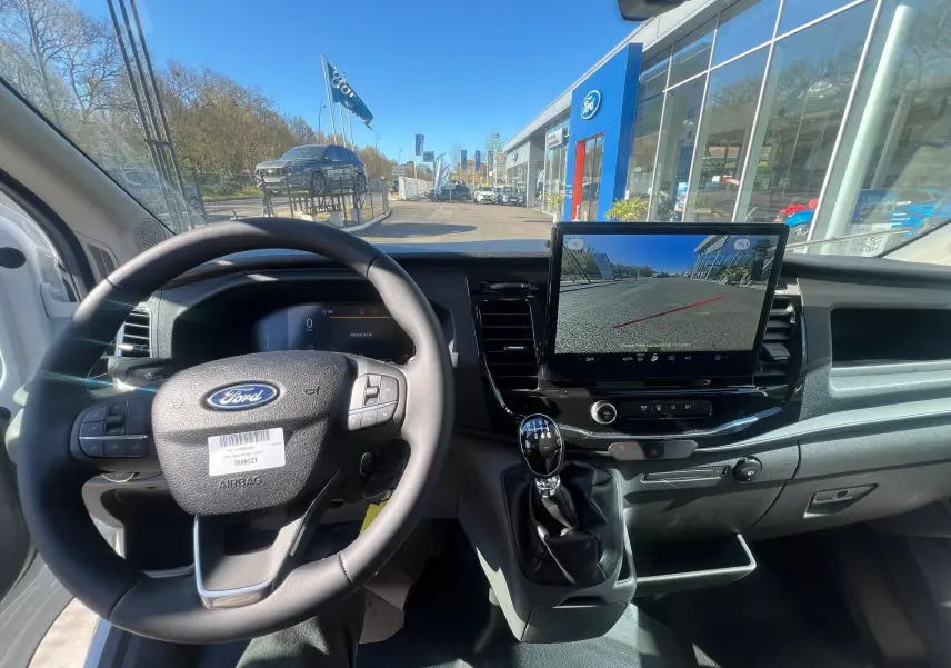 Vue intérieure du poste de conduite du Ford Transit blanc, avec volant, levier de vitesse manuel et écran tactile affichant la caméra de recul.