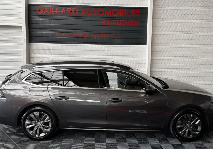 Profil droit d'une Peugeot 508 break gris foncé stationnée devant un garage avec sol damier noir et gris.