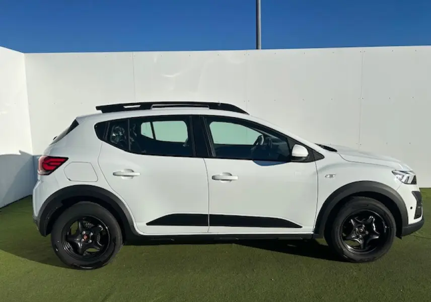 Vue de profil droit d'une Dacia Sandero Stepway blanche avec barres de toit noires et jantes foncées sur fond extérieur.