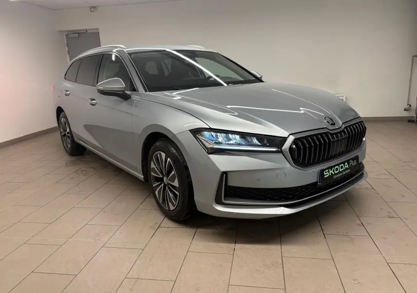 Skoda Superb Combi 2025 gris argent en 3/4 avant droit, avec calandre noire et phares LED allumés.