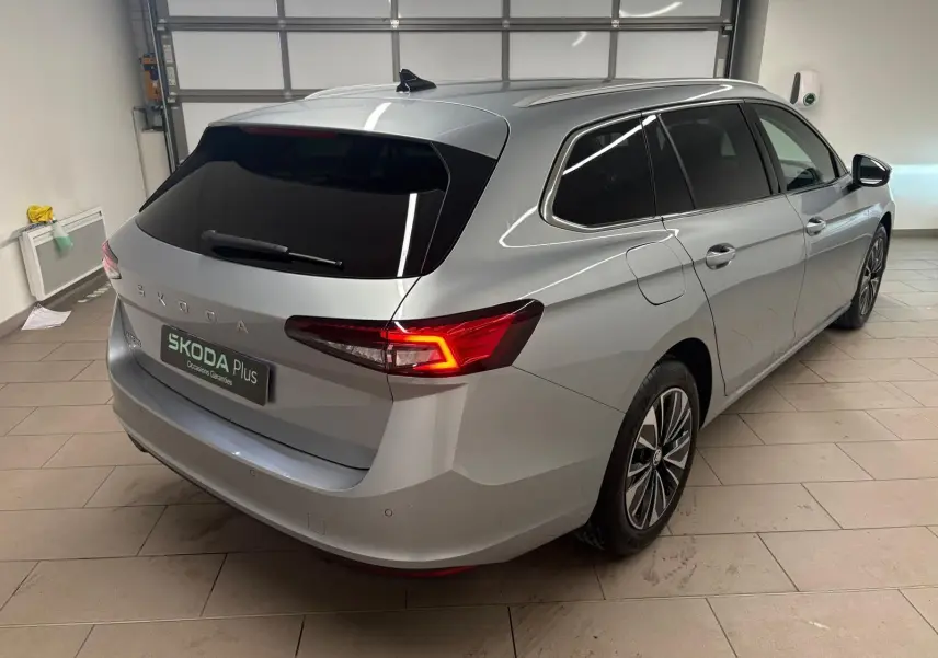Vue 3/4 arrière droite d'une Skoda Superb Combi gris argent 2025 avec feux arrière allumés dans un garage.