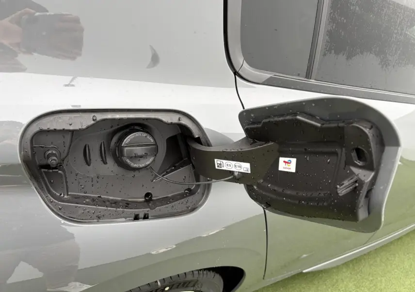 Trappe de carburant ouverte côté gauche d’une Peugeot 308 gris selenium avec bouchon essence visible sous la pluie.