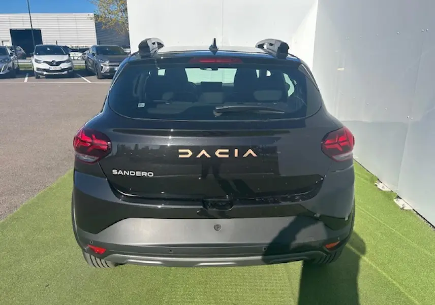 Vue arrière d'une Dacia Sandero III Stepway noir nacré avec logo Dacia brun cuivré et barres de toit visibles.