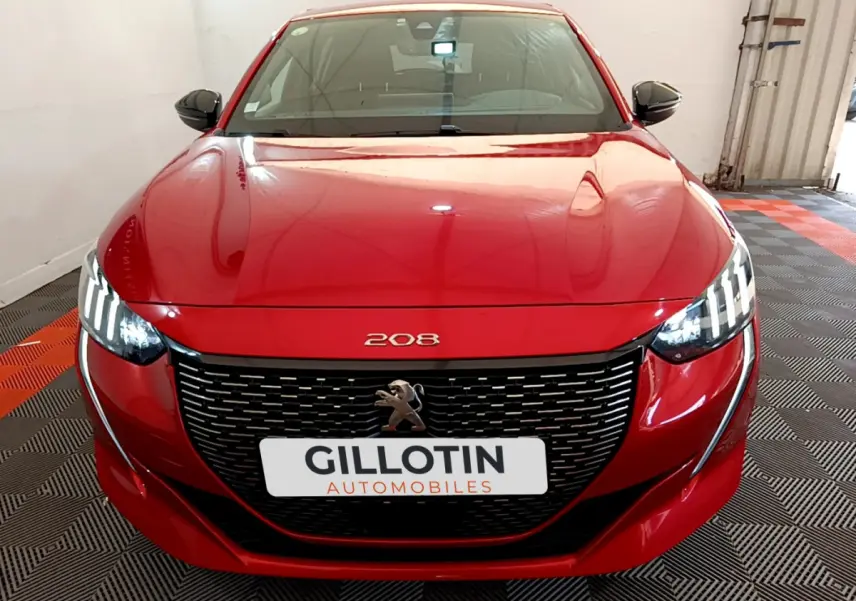 Peugeot 208 rouge vue de face, avec calandre noire et phares LED distinctifs allumés dans un garage.