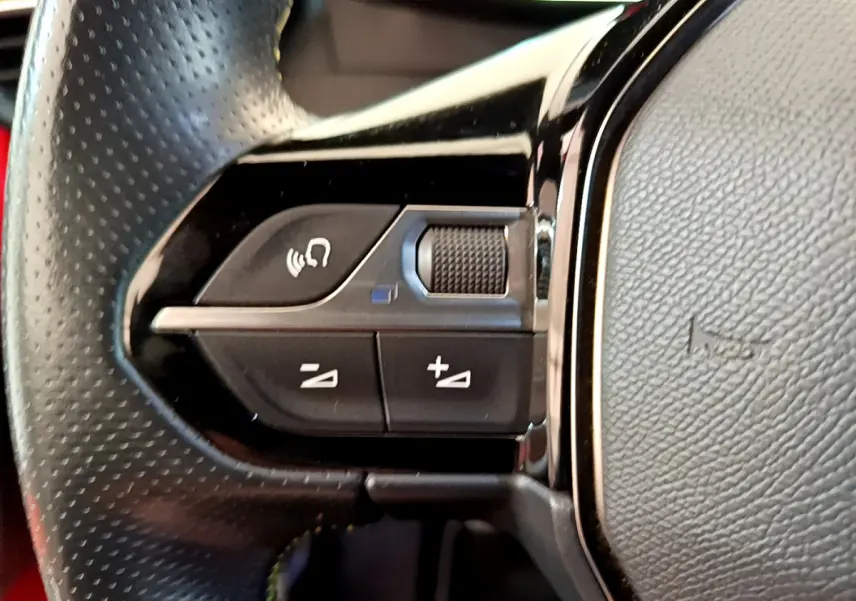 Gros plan sur les commandes gauche du volant en cuir perforé d'une Peugeot 208 rouge, version GT Line.