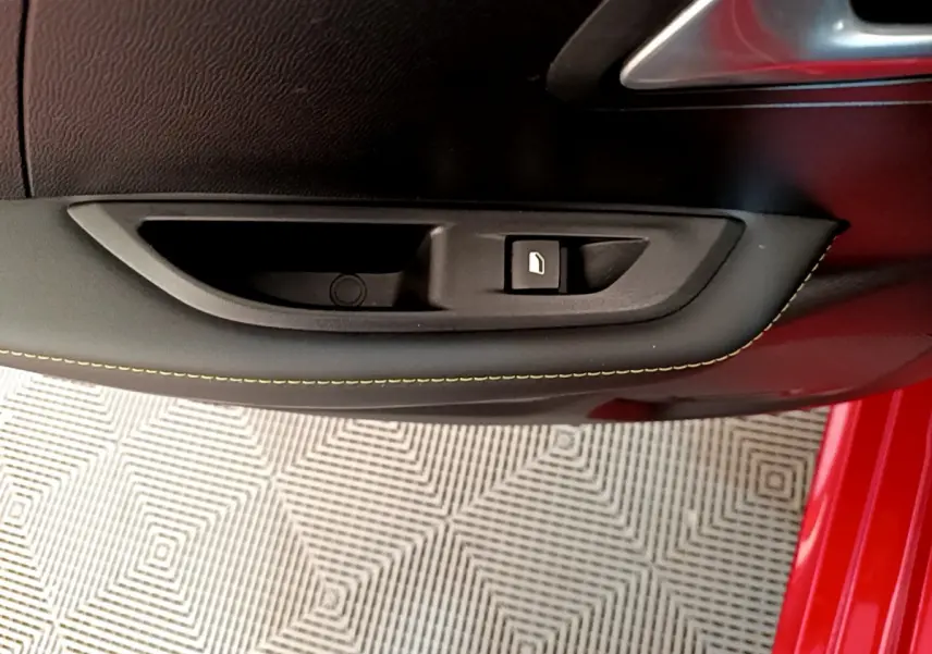 Gros plan sur la commande de lève-vitre côté gauche d'une Peugeot 208 rouge avec surpiqûres jaunes sur la portière.