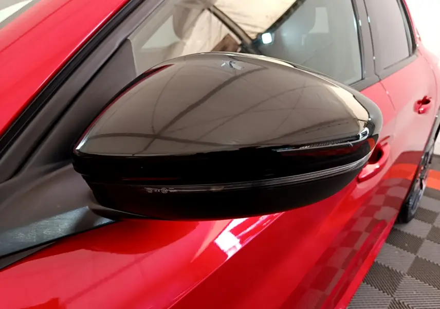 Rétroviseur noir brillant côté gauche d'une Peugeot 208 rouge GT Line vue en gros plan sur fond intérieur.