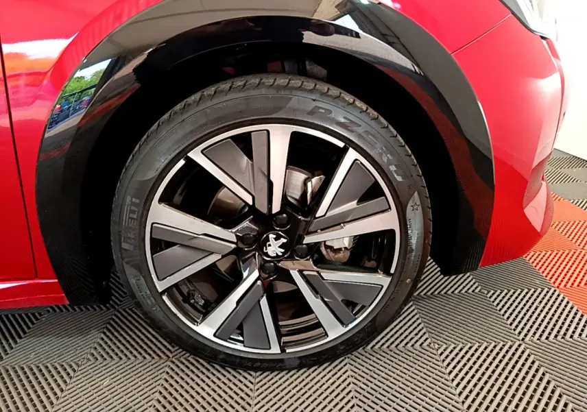Gros plan sur la roue avant gauche d'une Peugeot 208 rouge, jante alliage noire et argentée au design moderne.