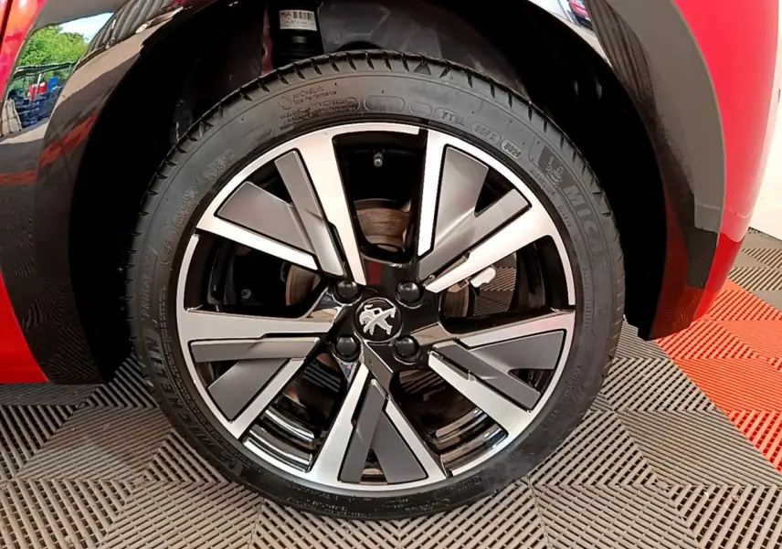 Gros plan sur la roue avant gauche d'une Peugeot 208 rouge avec jante alliage au design moderne et logo lion central.