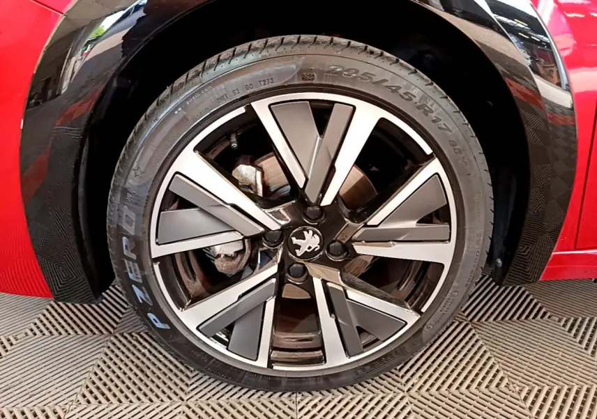 Gros plan sur la roue avant gauche de la Peugeot 208 rouge, jante alliage à design moderne et logo lion central.