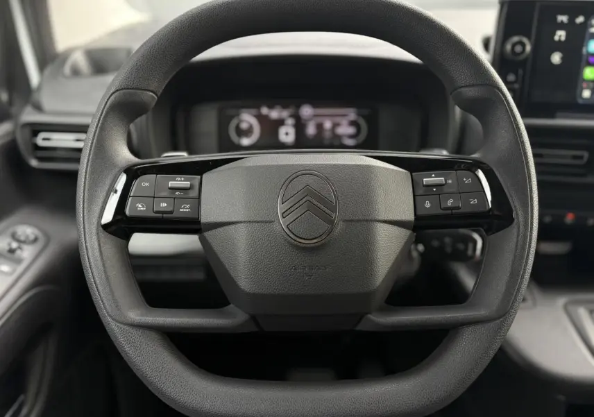 Vue rapprochée du volant multifonctions noir du Citroën Berlingo 2025 avec tableau de bord numérique en arrière-plan.