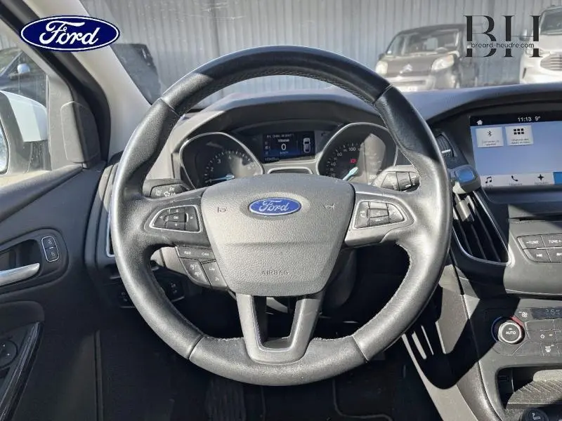 Vue intérieure centrée sur le volant cuir multifonction de la Ford Focus blanc glacier, avec tableau de bord et écran tactile visibles.