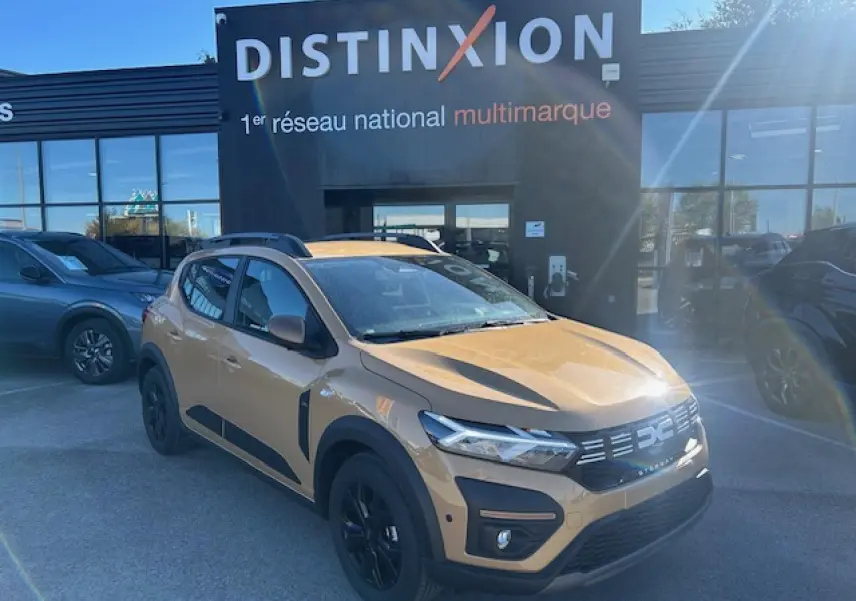 Dacia Sandero III Stepway beige Safari vue 3/4 avant droit, avec barres de toit et jantes noires.