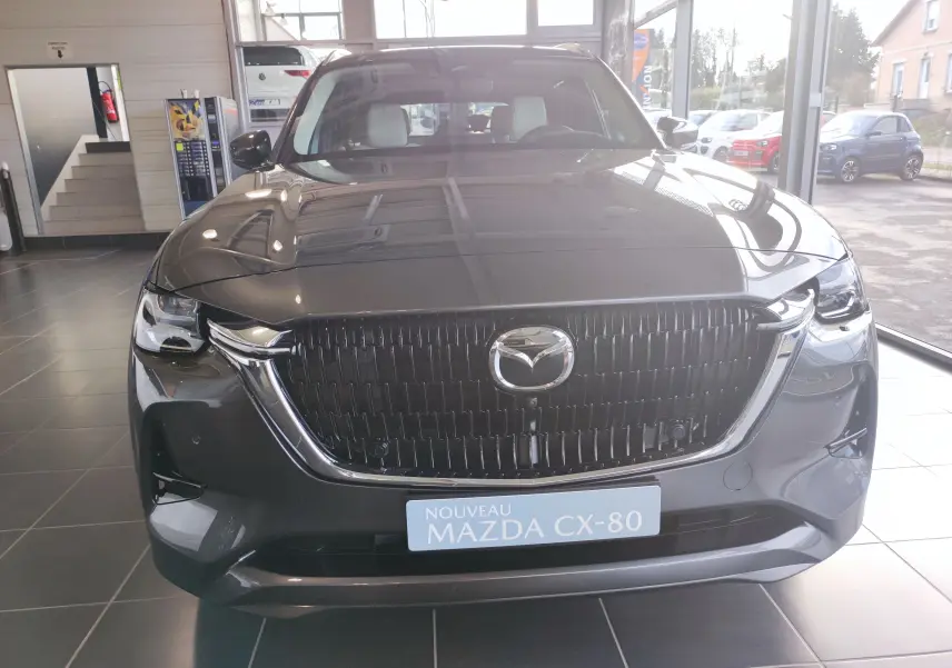 Vue frontale du Mazda CX-80 gris foncé 2024 en showroom, mettant en valeur sa calandre imposante et ses phares LED.