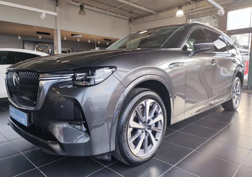 Vue 3/4 avant droit du Mazda CX-80 gris foncé 2024 en hybride rechargeable, avec calandre imposante et jantes alliage.