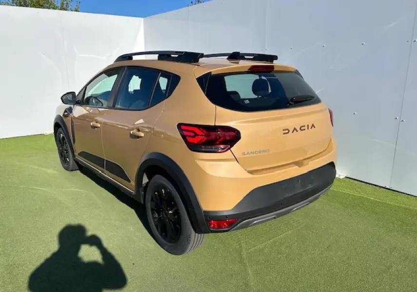 Vue 3/4 arrière droite d'une Dacia Sandero beige Safari avec barres de toit noires et jantes aluminium noires.