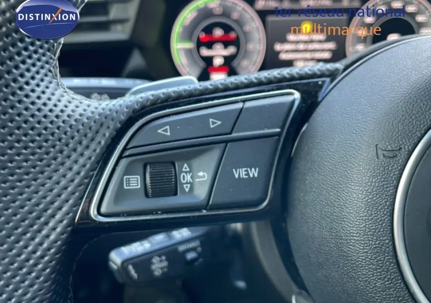 Gros plan sur les commandes multifonctions du volant perforé d'une Audi A3 Sportback blanc ibis, tableau de bord numérique en arrière-plan.