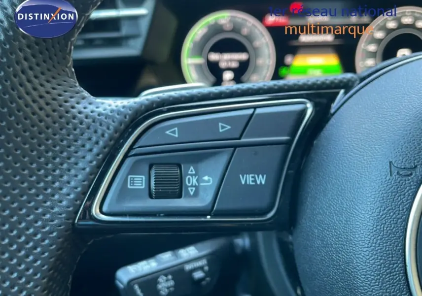 Gros plan sur les commandes gauche du volant perforé d’un Audi A3 Sportback 40 TFSIe noir Mito Metal, tableau de bord numérique en arrière-plan.