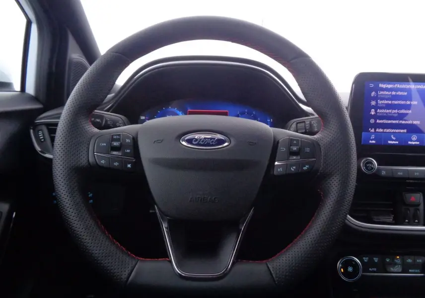 Vue rapprochée du volant perforé avec surpiqûres rouges du Ford Puma blanc, tableau de bord et écran tactile en arrière-plan.