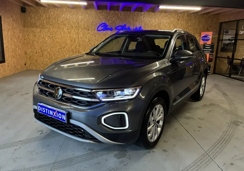 Volkswagen T-Roc gris 3/4 avant droit, phares allumés, calandre chromée et jantes alliage visibles en intérieur showroom.