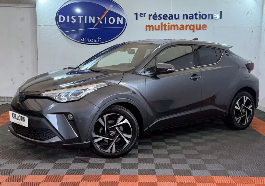 Toyota C-HR 1.8 Hybride gris foncé vu de profil avant droit avec jantes noires et logo Toyota visible.