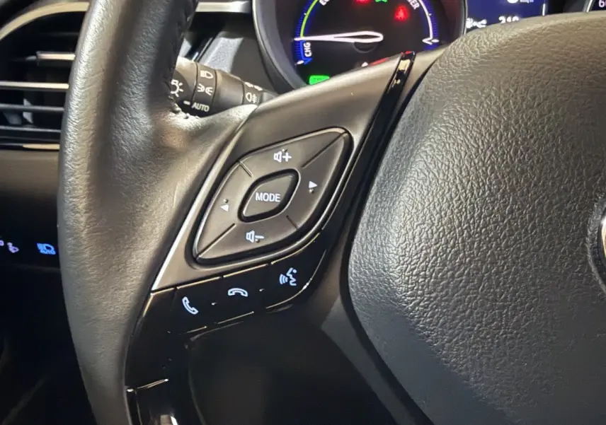 Gros plan sur le volant cuir noir du Toyota C-HR 1.8 Hybride 2022 avec commandes multimédia et tableau de bord lumineux.