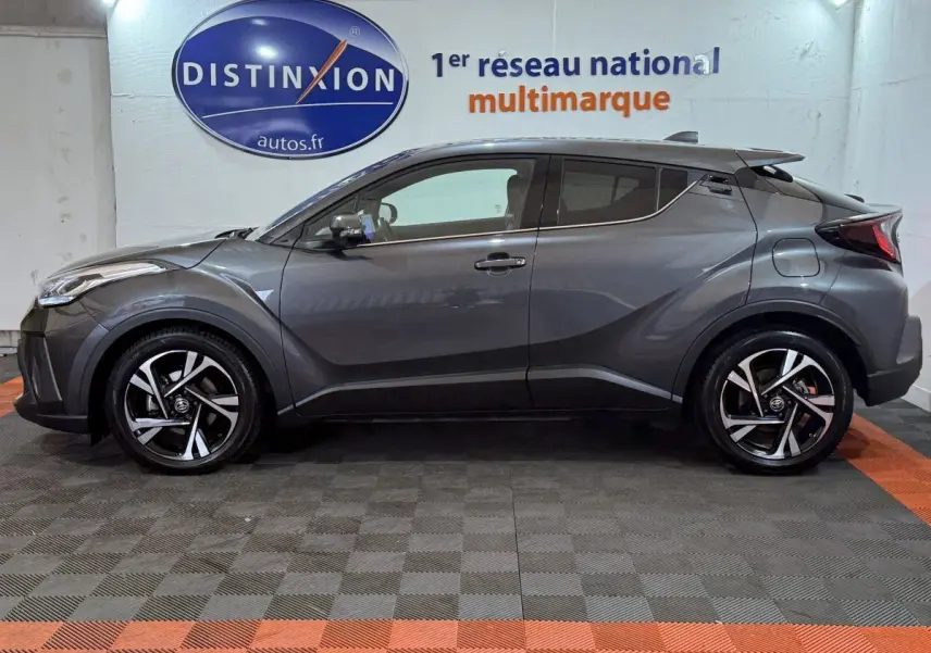 Profil latéral droit d'un Toyota C-HR 1.8 Hybride gris foncé avec jantes alliage et toit noir en intérieur showroom.