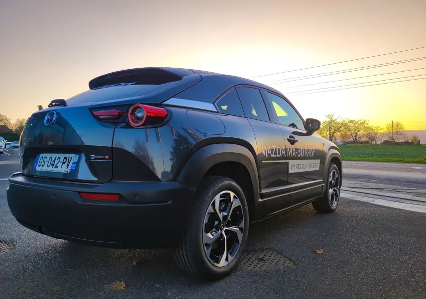 Vue 3/4 arrière droite d'un Mazda MX-30 gris foncé hybride rechargeable avec marquage latéral au coucher du soleil.