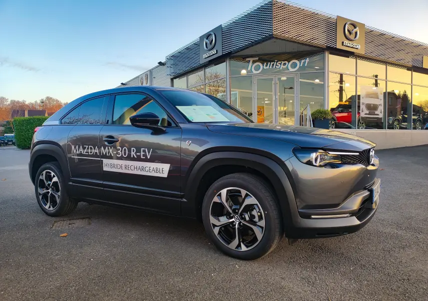 Mazda MX-30 hybride rechargeable gris foncé vue 3/4 avant droit devant un concessionnaire Mazda.