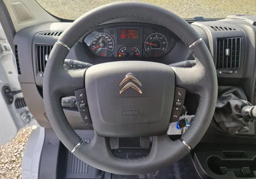 Vue intérieure centrée sur le volant noir perforé du Citroën Jumper blanc 2023 avec tableau de bord et levier de vitesses visible.