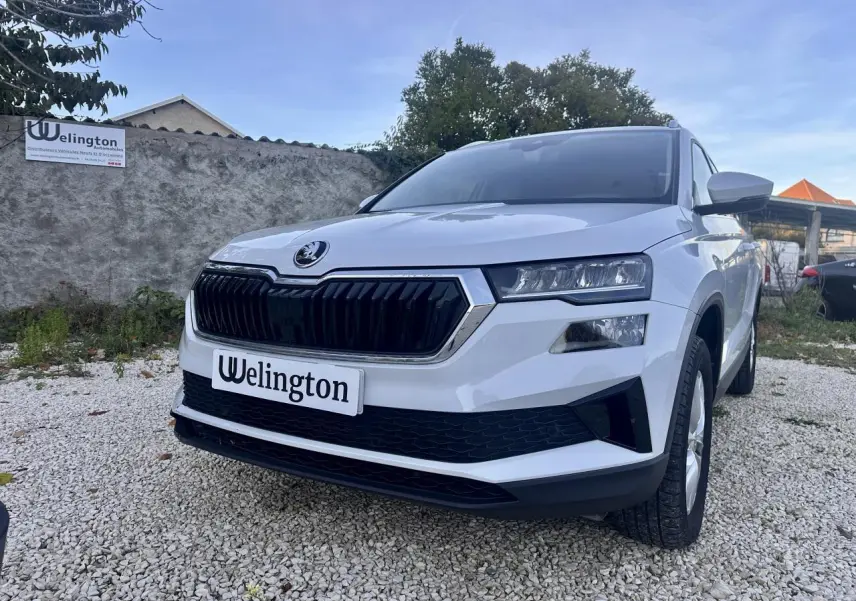 Vue 3/4 avant d'un Skoda Karoq blanc 2024 avec calandre noire et phares LED, sur un sol gravillonné.