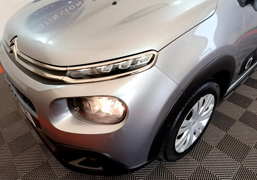 Vue 3/4 avant droit d'une Citroën C3 gris clair avec phares allumés et jantes acier au design spécifique.