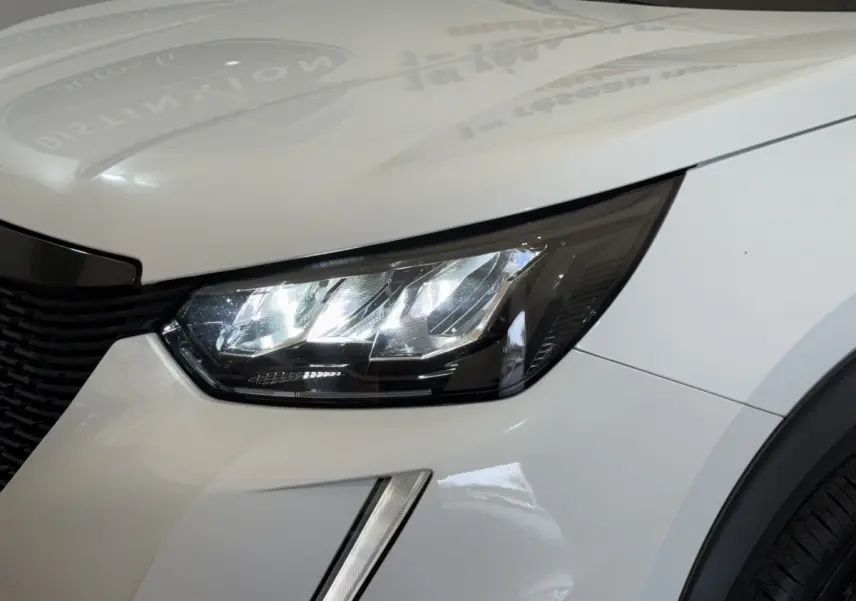 Gros plan sur le phare avant droit LED du Peugeot 2008 blanc, mettant en valeur la calandre et la carrosserie lisse.