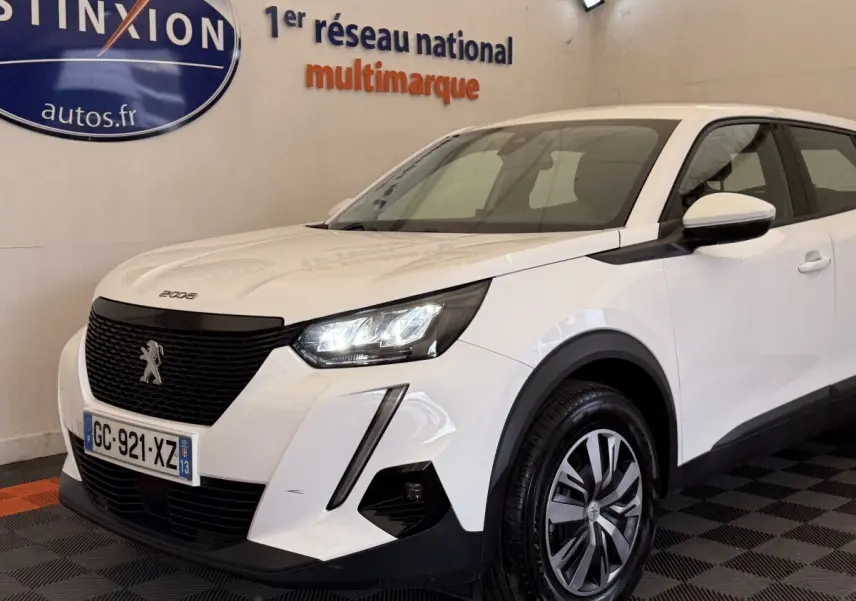 Peugeot 2008 blanc vue 3/4 avant droit, avec calandre noire et feux LED allumés en intérieur showroom.