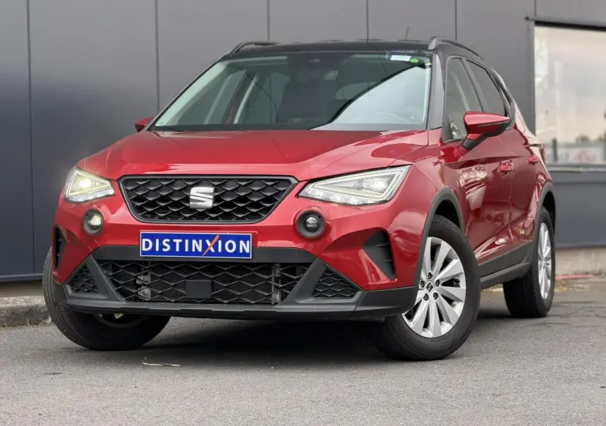 Vue 3/4 avant d'un SEAT Arona rouge Désir avec toit noir et jantes alliage, stationné devant un mur gris.