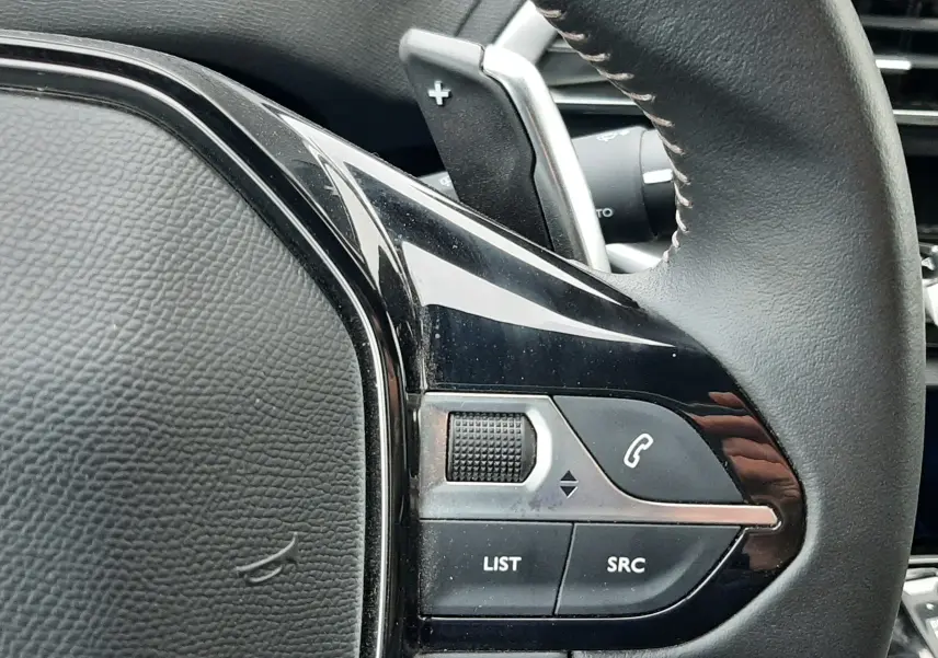 Gros plan sur la partie gauche du volant noir du Peugeot 3008 Hybrid 136 e-DCS6 Allure Pack, avec commandes audio et palettes de changement de vitesse.