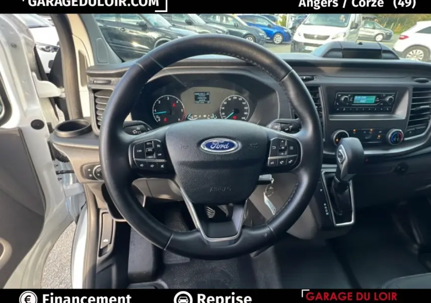 Vue intérieure centrée sur le volant multifonctions en cuir noir du Ford Transit Custom blanc, avec tableau de bord et levier automatique.