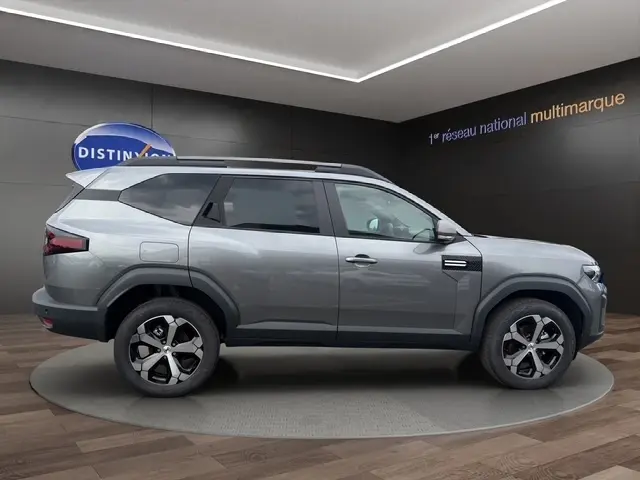 Vue de profil côté droit du Dacia Bigster gris foncé avec jantes alliage et barres de toit noires en showroom.