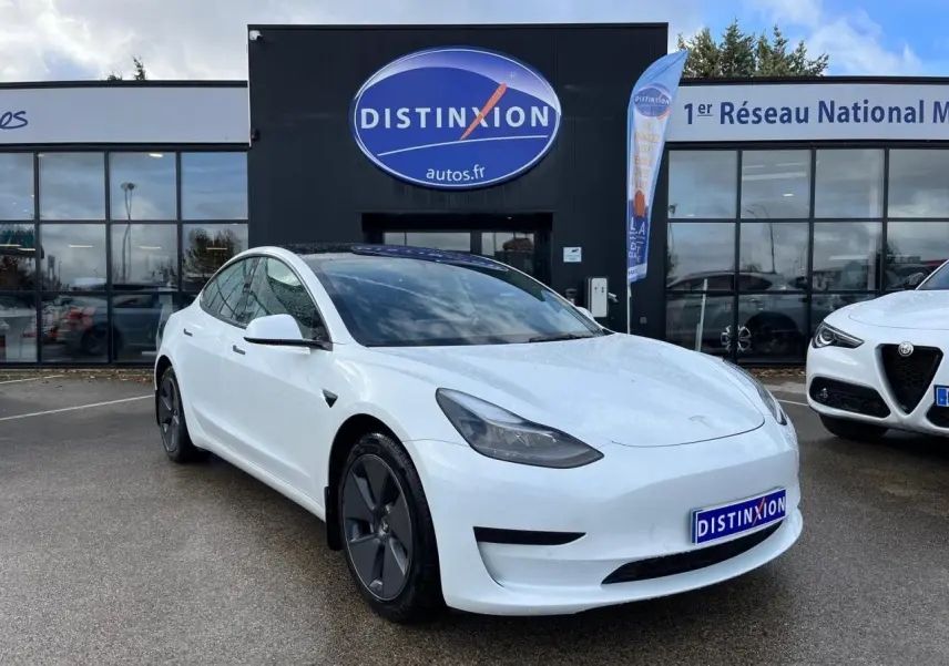 Tesla Model 3 blanc en 3/4 avant droit, avec toit panoramique en verre et jantes aérodynamiques noires.