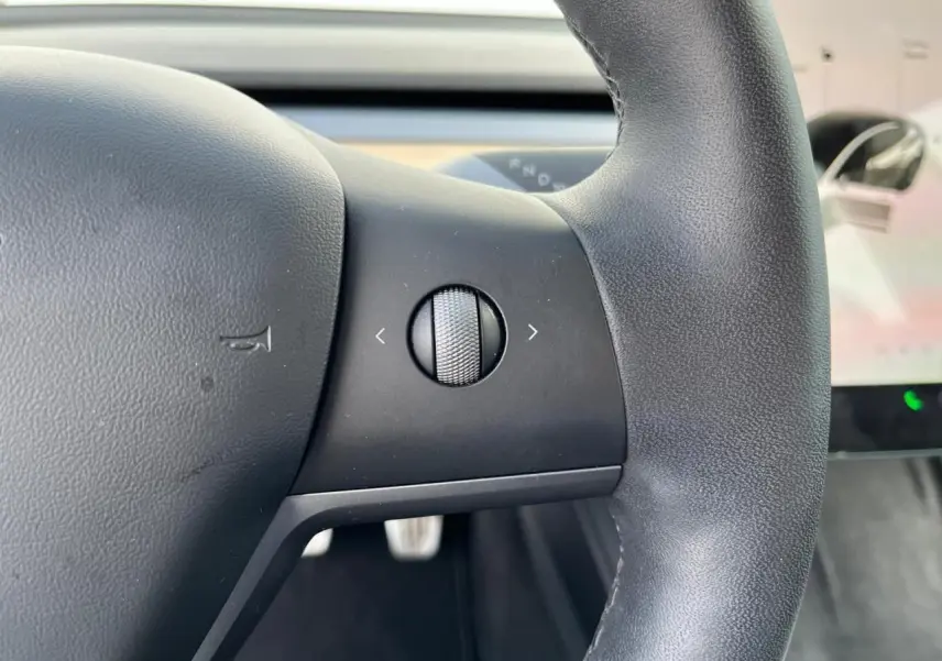 Gros plan sur la molette de commande à gauche du volant noir d'une Tesla Model 3 blanche, intérieur moderne visible.