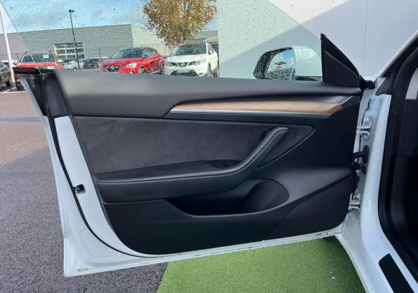 Porte avant gauche ouverte d'une Tesla Model 3 blanche, intérieur noir avec bandeau décoratif en bois.