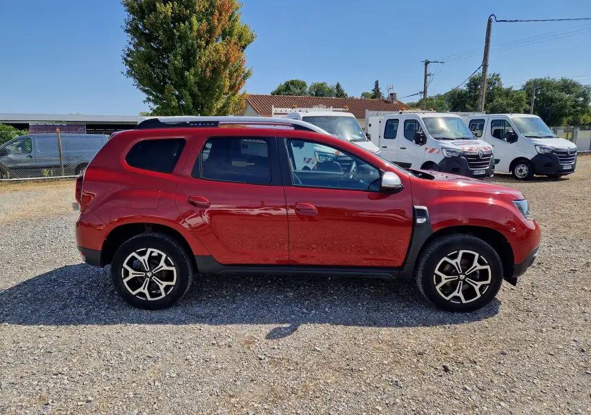 Profil droit d'un Dacia Duster rouge fusion 2021 avec jantes alliage noires et barres de toit argentées.