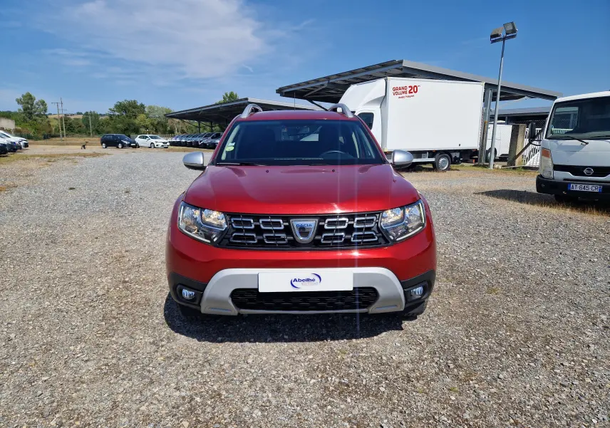 Vue de face d'un Dacia Duster 2021 rouge fusion avec calandre noire et détails argentés sur un sol gravillonné.